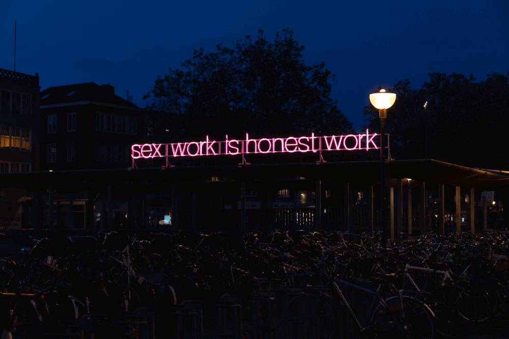A photograph of a dimly-lit building at night. On top of the building's roof, a pink neon sign reads "sex work is honest work." The neon casts a dim pink light on the many rows of bikes in front of the building.
