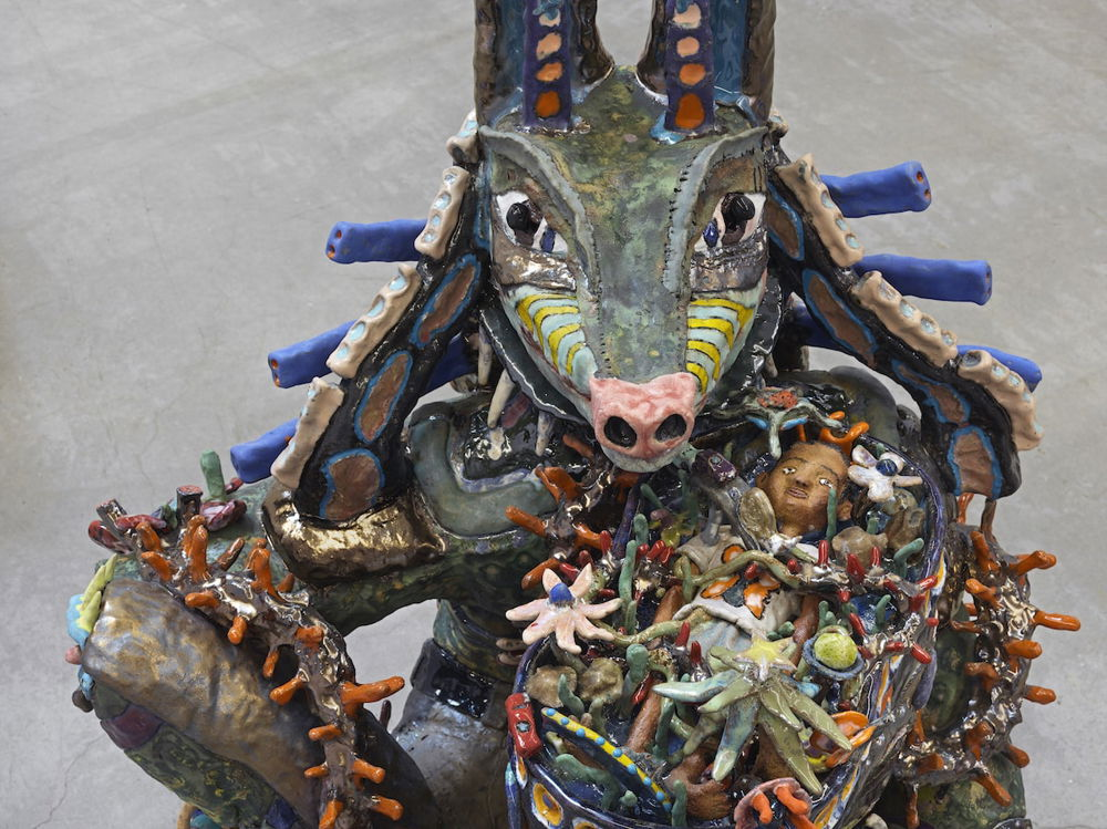 A sculpture of an anthropomorphized animal, with a snout and long ears. The animal is painted a greenish grey, with decorative dashes and dots. He is covered in ceramic floral shaped objects that also obscure a small boy the animal holds in a basket.