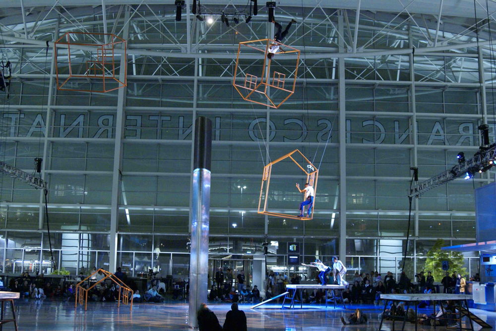 Three orange armatures resembling houses are suspended by cables in the cavernous glass lobby of the San Francisco International Airport. Two aerial dancers perform within the suspended structures, while two more perform on the ground below, reaching toward another orange house planted on the floor that appears to be halfway submerged.