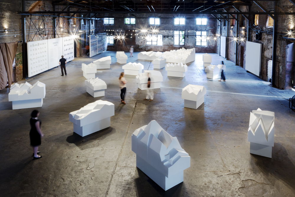 Sixteen large, white, foam models of roof architecture are spotlit in a large warehouse-like exhibition space.
