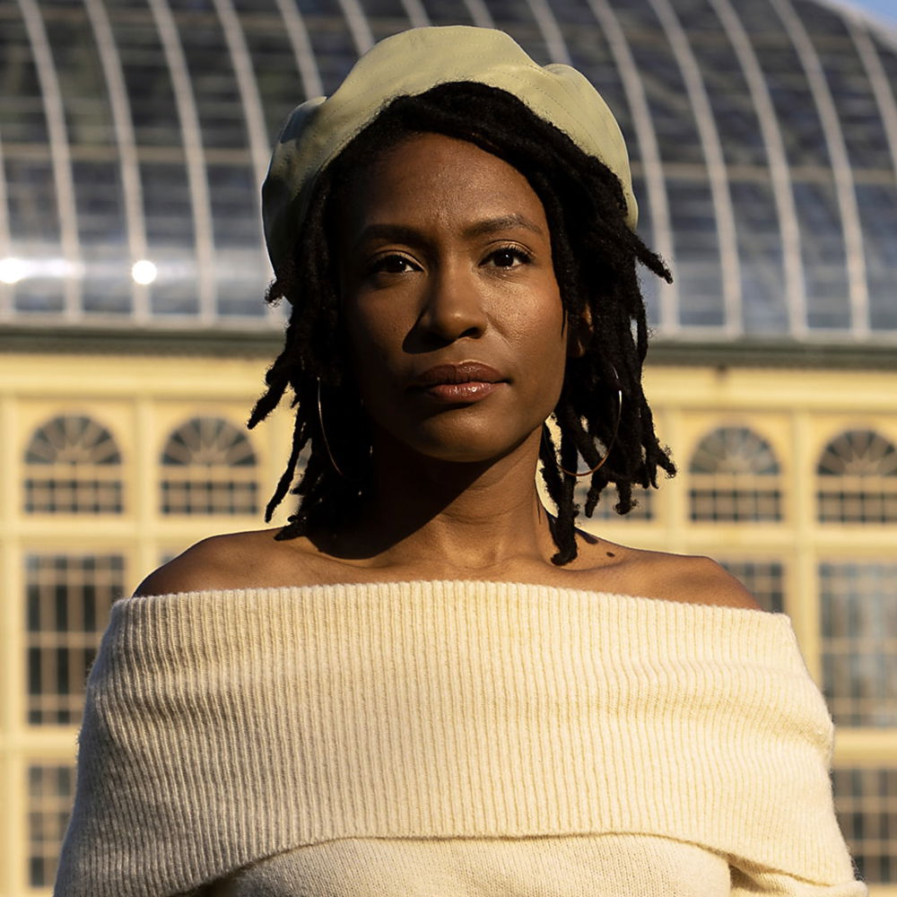 A brown-skinned Black woman with shoulder-length locs standing in front of a domed plant conservatory with shrubs and plants in the background. She is wearing a sage-green leather beret and an off-the-shoulder cream-colored sweater.