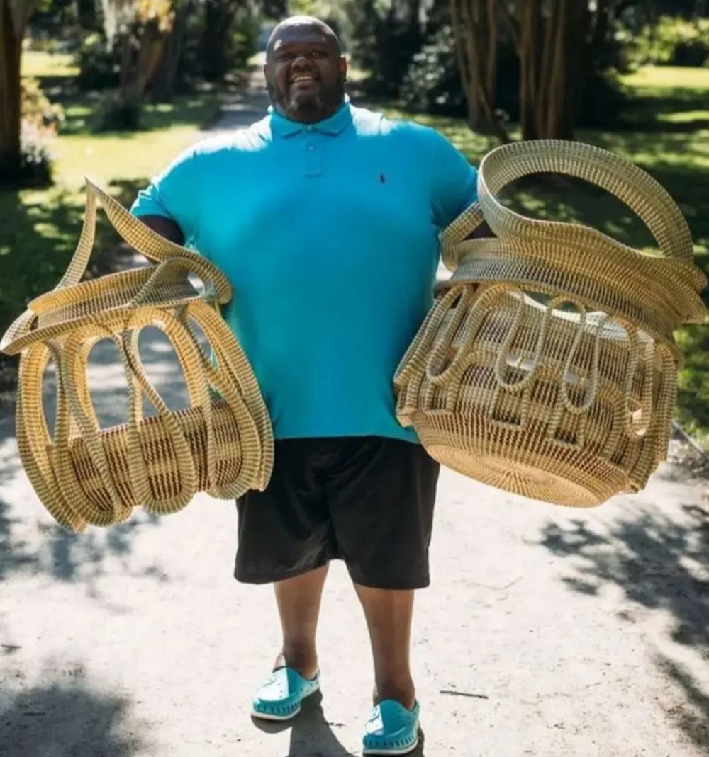 [ID: A photograph of the artist, Corey Alston, standing in a park holding up two large woven baskets constructed with wavy organic lines and shapes. Alston wears a bright-teal polo shirt, black shorts, and coordinated teal shoes.]