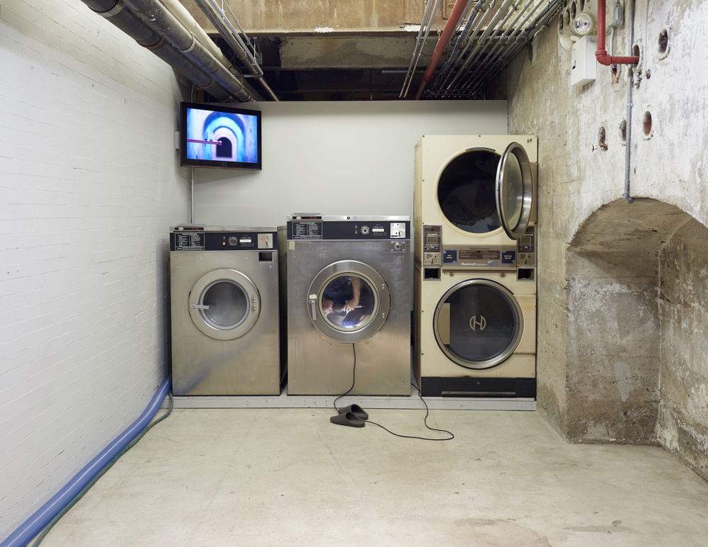 A view of laundry machines and a television monitor mounted high on the wall. Sasamoto spins inside a modified washing machine, holding the microphone as she reads from a book. The monitor screens the view from inside the washer.
