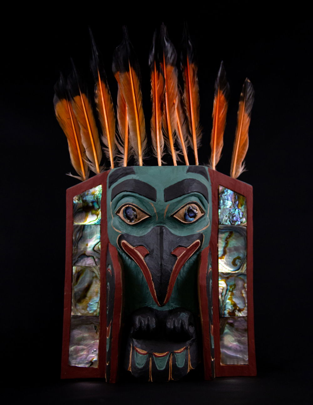 A raven headdress frontlet traditionally put on the front of an ermine skin headdress. It represents a raven, one of the main moieties in Northwest coast culture. The raven’s head, claws, tails, and wings are compacted together in the center and surrounded by abalone inlay on either side, with orange and black-tipped flicker feathers attached to the top of the frontlet.]