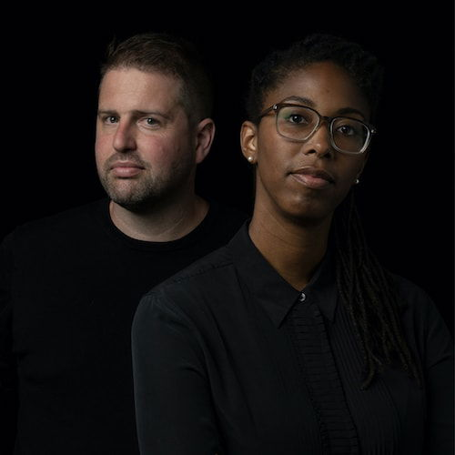 A headshot of two people. Jennifer, an African-American woman with glasses and long natural locs, wears a black shirt and looks directly at the camera with her head tilted to the side. Tom, a white male with brown hair and a subtle beard, wears a black shirt and looks directly at the camera. Both are standing in front of a black background.
