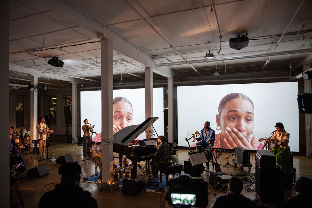 Musicians perform in a space in front of a video installation. Both of the two screens behind the musicians project the same image, a Black woman placing a hand over her mouth.