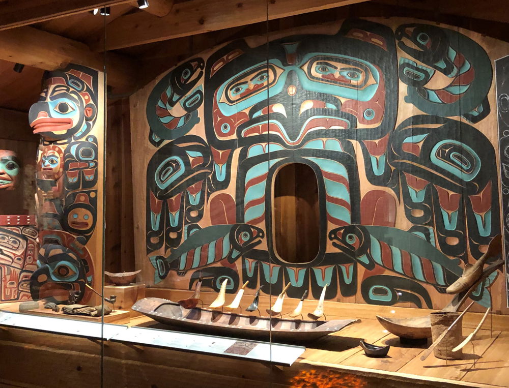 This house screen on display in the Alaska State Museum in Juneau features a raven and four dog salmon in the traditional Northwest coast design style. It is a symmetrical panel painted in the traditional colors of blue green, red, and black. The four dark bluish green dog salmon are positioned in the four corners and the raven is centered with outspread wings. Two dog salmon jumping at the top are quite happy.