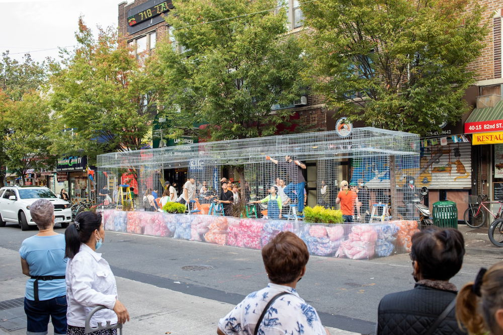 Photo of a team of people installing a public seating area in Jackson Heights, Queens, as neighborhood residents look on. The structure is built in the street in front of a row of businesses and is made of a wire frame and transparent plastic, with colorful bags of insulation visible inside.