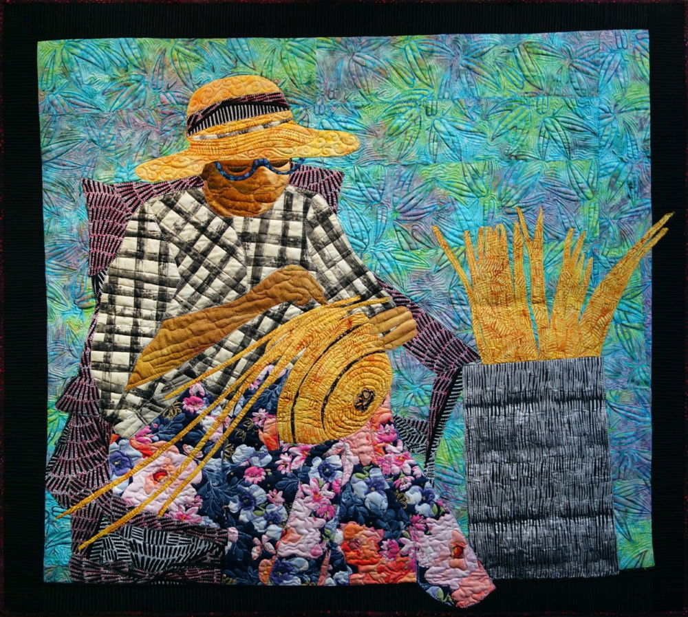 A quilt of many textures, colors, and patterns depicting a Black woman weaving a basket in a wicker chair. She sits on a field of turquoise, green, and purple fabric.