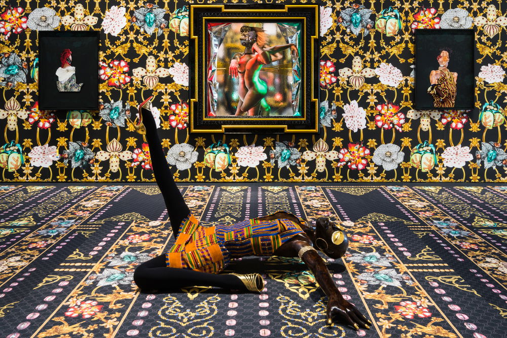 A sculpture of a Black woman lays on the floor, one leg straight up in the air, the other bent back at her side. Her arms are fanned out, head bent back, gold disc eyes closed. In the background, there are three portraits depicting various figures. The floor, walls, and clothing are a cacophony of floral and geometric patterns.