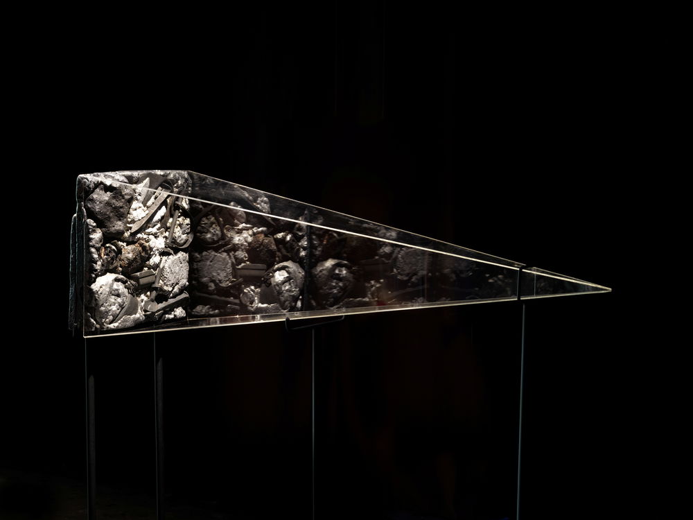 A pyramid vitrine lays on its side shrouded in darkness. Inside the vitrine is an illuminated collage of railroad nails, shackles, and various metals.