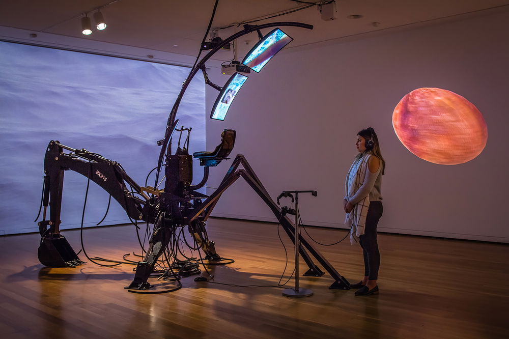 A refurbished interstellar backhoe outfitted with a bucket loader, three LCD monitors, multiple pairs of headphones, and long, spindly legs sits in the center of the gallery. The wall to the left displays a video projection of a 3D rendered ocean and the wall to the right displays a projected red orb.