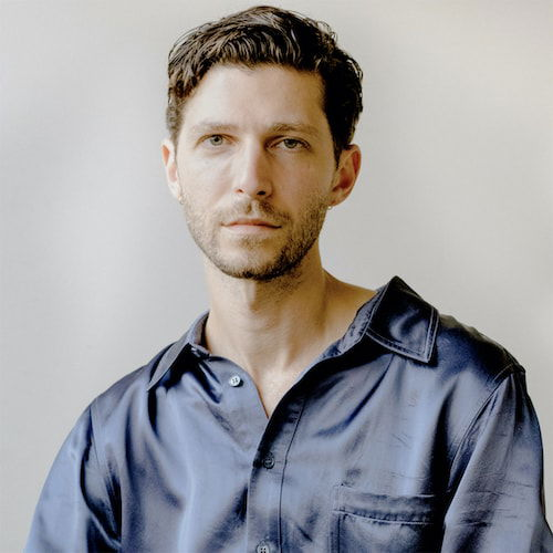 A portrait of a man with a pale complexion sitting in front of a light grey background. He has short, cropped brown hair, and wears a blue silk button-down shirt.