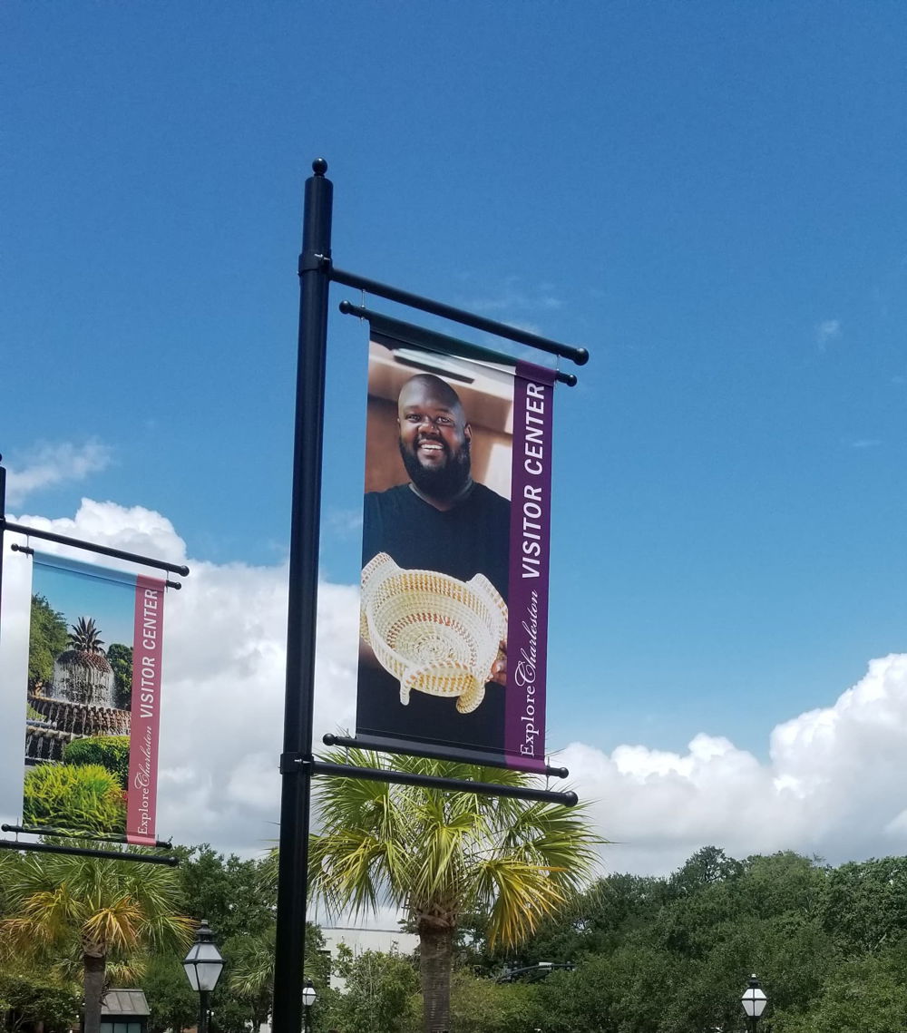 [ID: A street banner featuring a photograph of the artist Corey Alston. He smiles and holds a beige-colored woven basket. To the right of the photo is a bar of vertical white text on a purple background that reads, "Explore Charleston Visitor Center."]