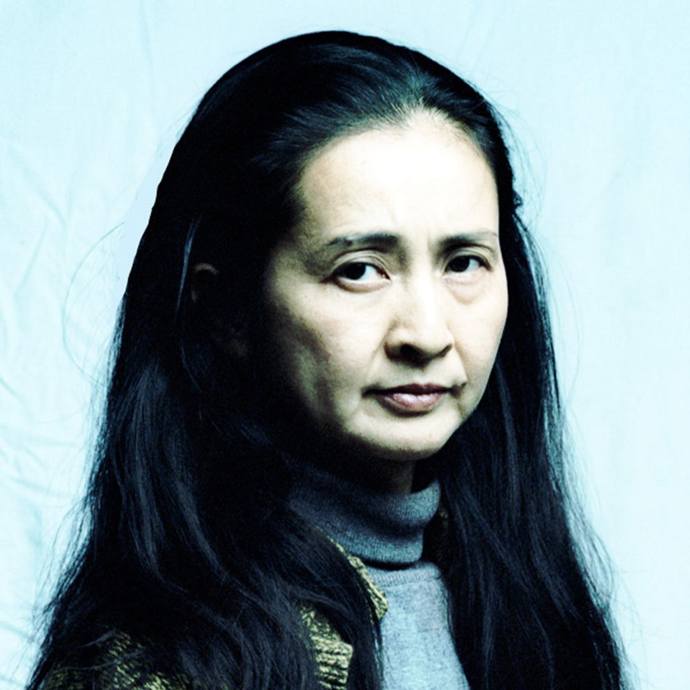 A headshot of a Japanese woman wearing a gray turtleneck sweater and green cardigan. She looks over her right shoulder to stare directly at the viewer with brown eyes. She has long, black hair and is slightly smiling.