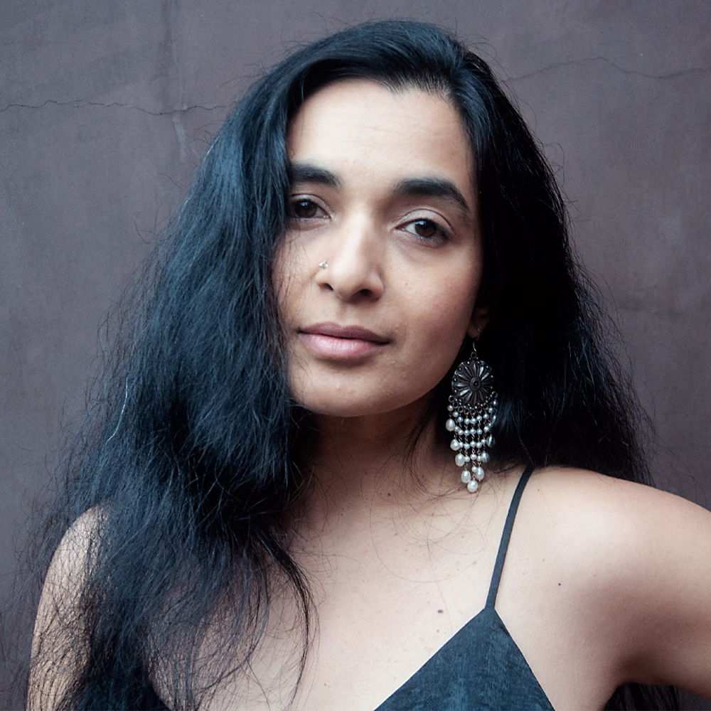 A portrait of a South Asian artist with brown skin, dark brown eyes, and long dark hair falling on the right side of her face. She's looking straight into the camera, posed in front of a stucco wall. She is wearing a black silk blouse with spaghetti straps, and on her left ear is a hanging pearl earring from India.
