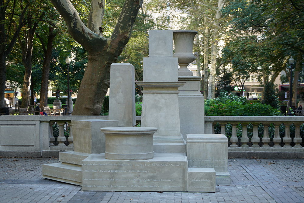 Photograph of a sculptural assemblage of nine monument pedestals without any statues on them sitting in front of a balustrade of a similar color in a city square in Philadelphia. A band of lettering at the base of the sculpture reads: "On this site there could be a statue to Jarena Lee, Sarah Allen, Notike..."
