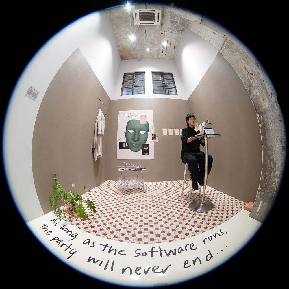 Viewed through a fisheye lens, a Chinese man sits at a table in a small room with brown walls and a tiled floor. A bathrobe and posters hang on the wall, and a plant and bar cart sit on the floor. In the foreground, a phrase written in marker reads “as long as the software runs, the party will never end...”