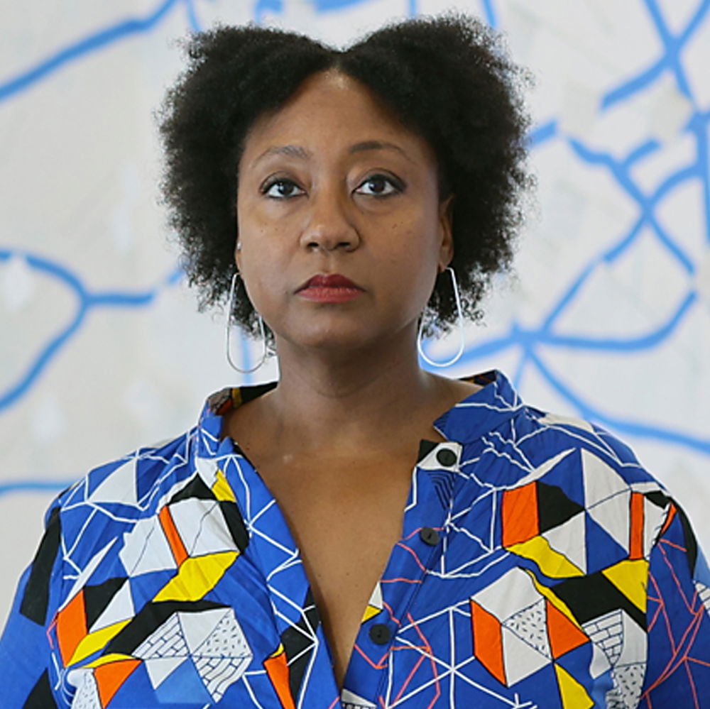 A Black woman with short hair looks out beyond the camera. She wears tear drop–shaped wire earrings, a colorful patterned shirt, and poses alongside a sculpture and mural.