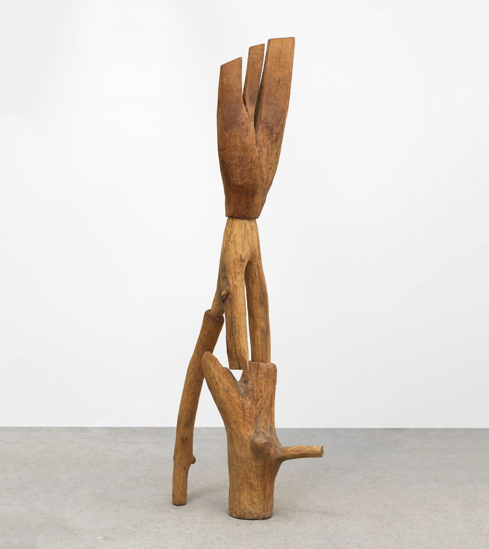 Photo of a sculpture formed from several pieces of wood in which the grain and natural shapes are still visible. The pieces are balanced together into a tower evoking a group of acrobats.