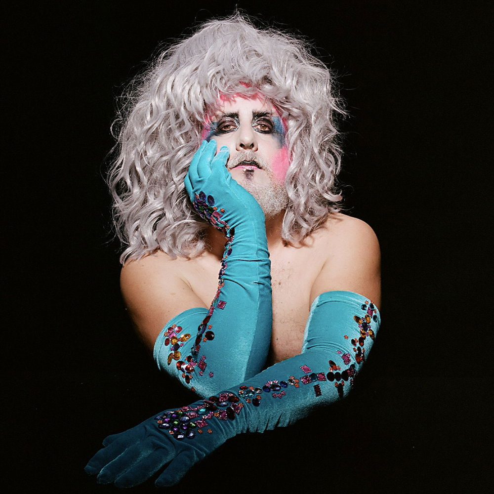 A portrait of a person wearing pale makeup accented by bright pink and turquoise around the eyes. He has a gray beard and face-framing wavy gray hair that reaches his bare shoulders. He wears sequined arm-length turquoise gloves and leans his head on one hand against a dark backdrop.