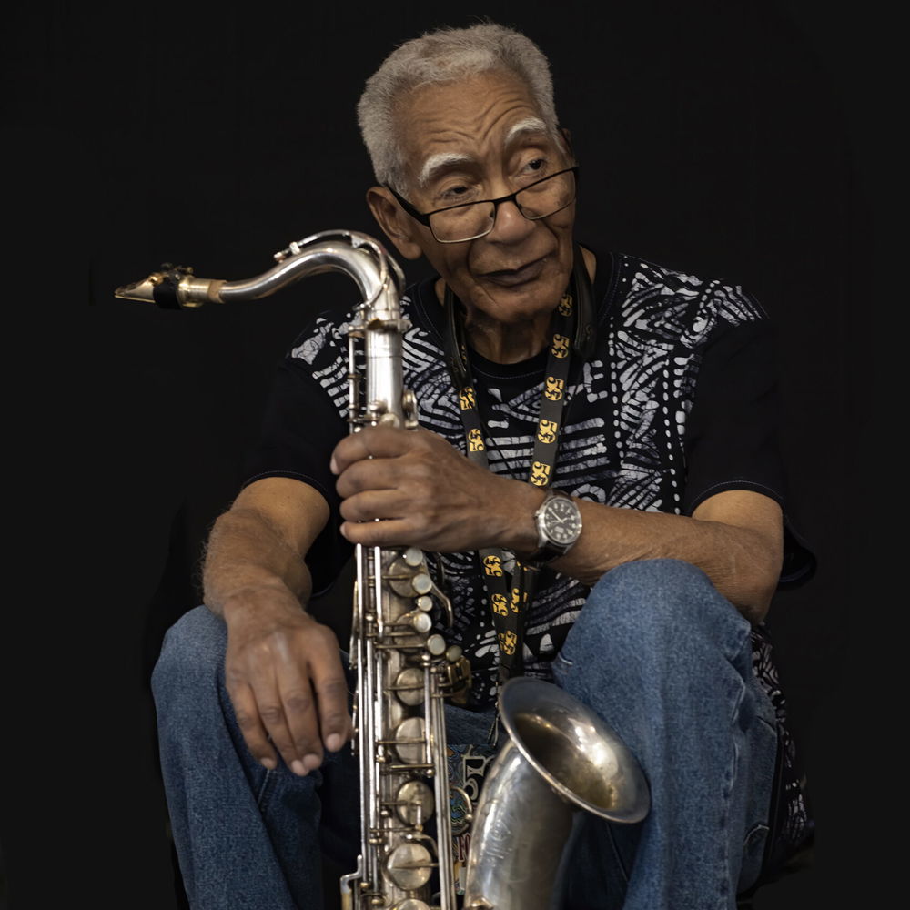 Edward "Kidd," a Black man with gray hair, a printed t-shirt, jeans, and his glasses low on his nose, sits holding his saxophone.