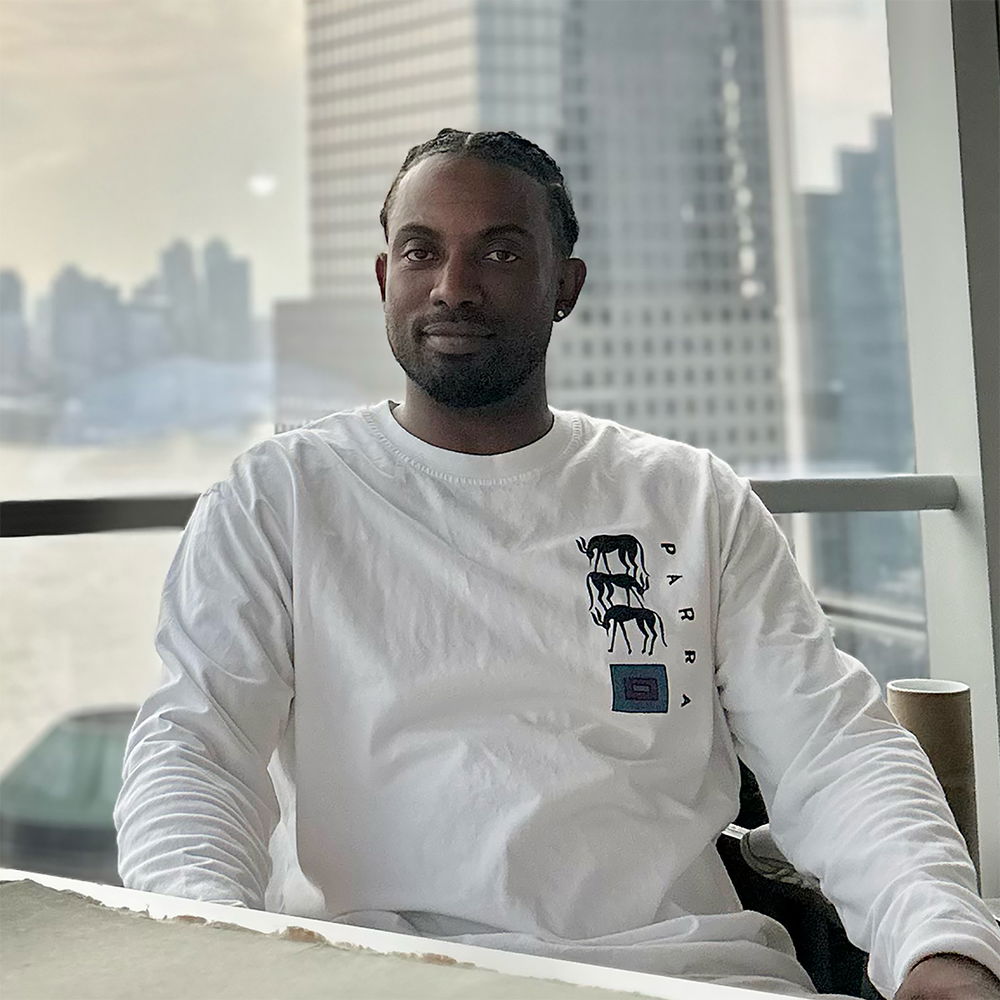 A Black man in a white long-sleeved shirt sits at a desk in an office space with a cityscape visible through the large glass window behind him.