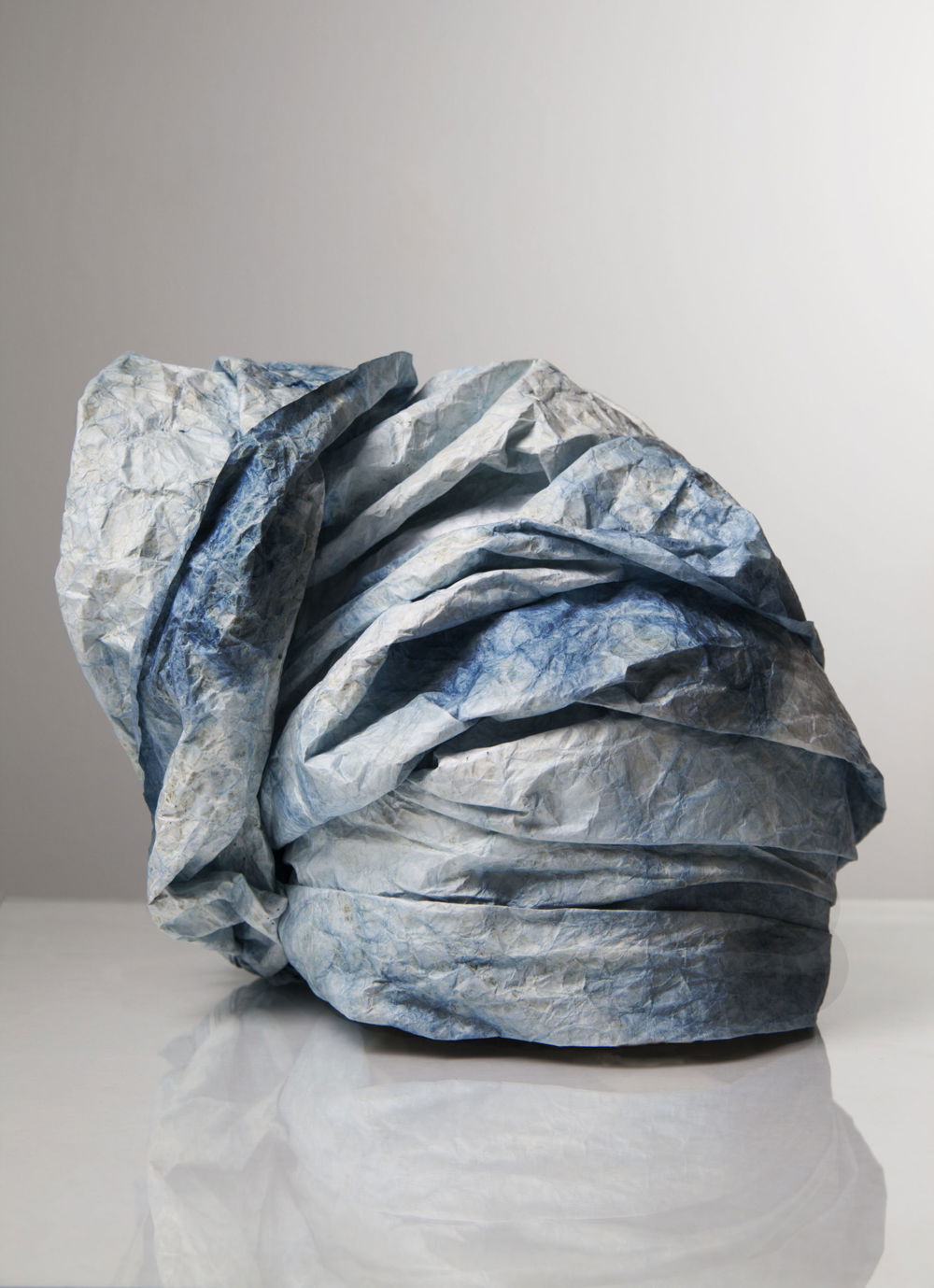 Photo of a piece of blue-dyed fabric, wound and folded around itself to form a sinuous, crownlike shape reminiscent of a Nigerian gele or head tie worn by Yoruba women. The dye is uneven and darker in some spots than others, showing a gently wrinkled texture.