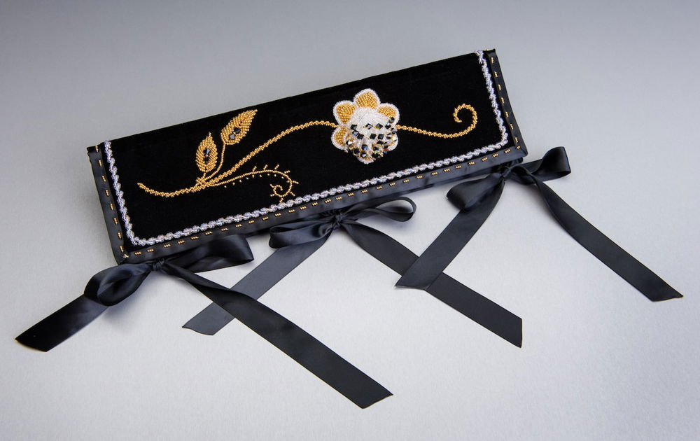 Golden and white beaded embroidery in the shape of a small flower and swirling stem on a rectangular piece of black fabric. Black ribbon frames the object and three delicate, black bows run along the bottom.