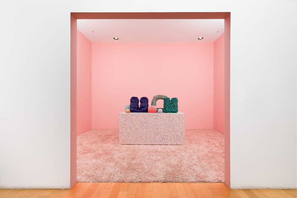 Photograph of an entryway that opens into a baby-pink colored room. At the center of the room, a pink rectangular table with silver detailing. Seven ceramic structures — all of different shapes, sizes and colors — rest artfully.