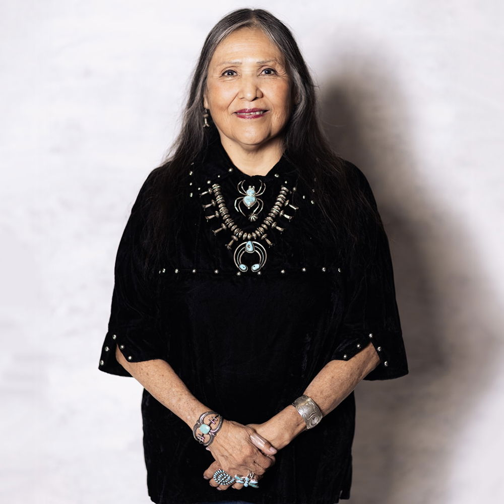A woman with shoulder length black and gray hair smiles at the camera with her hands clasped in front of her. She is wearing a black traditional Navajo outfit and traditional turquoise and silver jewelry, including bracelets, rings, and necklaces.