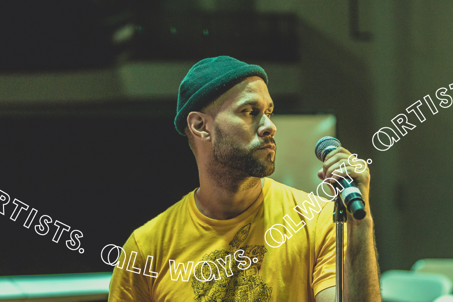 A Latinx man with a short beard wearing a yellow t-shirt and a black beanie holding a microphone on a stand.