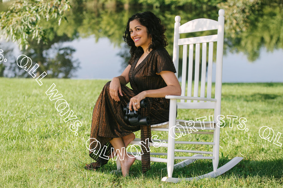 A South Asian Filipina woman woman with shoulder-length dark brown hair sits in a white rocking chair in front of a lake. She is barefoot and birdwatching, holding binoculars and wearing a metallic ankle-length dress.