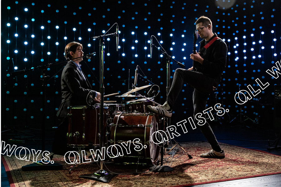 Two musicians are performing on a small stage, one seated at a drum kit and one standing with a guitar. They are lit by overhead spotlights that create a striking, moody atmosphere. The background is adorned with an expansive wall of LED lights, arranged in a grid of dots, which glow in a gradient, shifting from green on the left to blue and purple on the right. Microphones, stands, and other equipment are visible. The overall mood is vibrant and immersive, blending live music with a visually compelling light display.