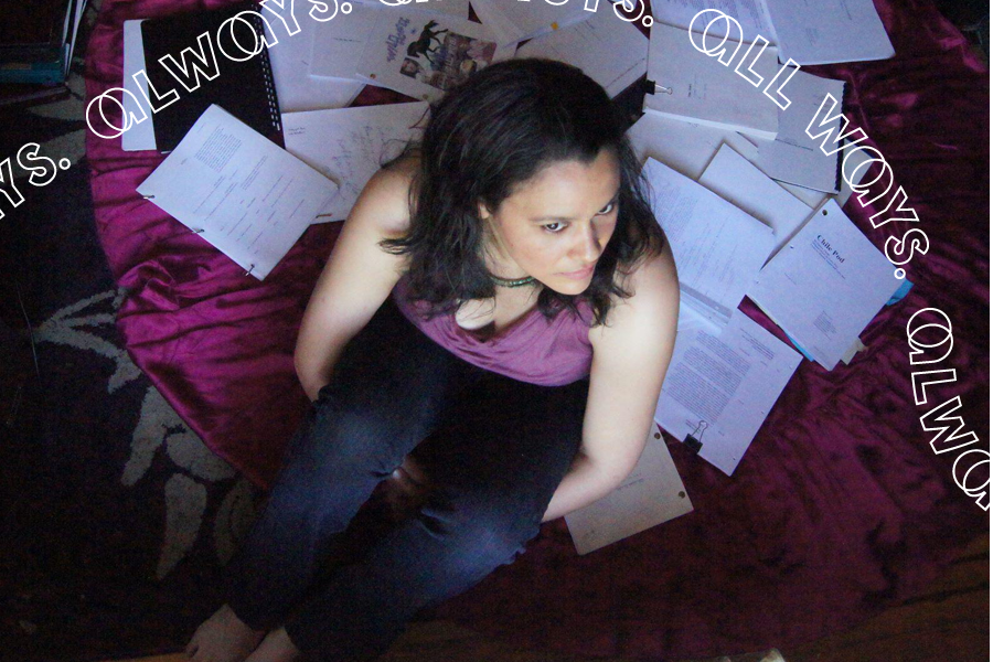 A view from above of woman sitting on a maroon cushion. Behind her is a wide array of play scripts printed on white paper.