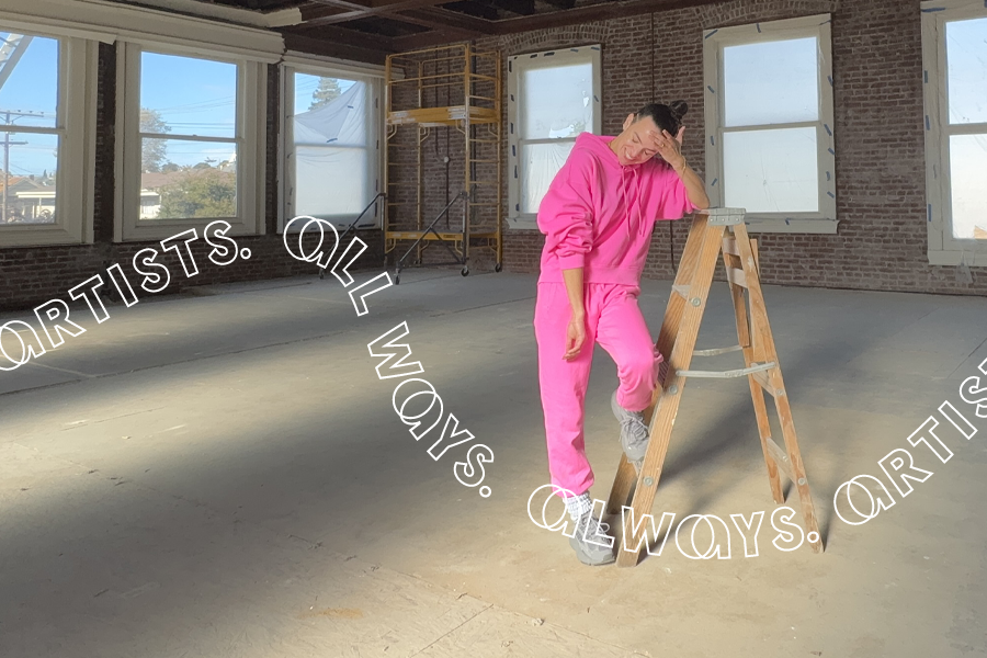 A portrait of a figure in a light-filled, and otherwise empty, industrial brick building. Wearing a neon pink sweatsuit, she laughs and leans on a wooden ladder.