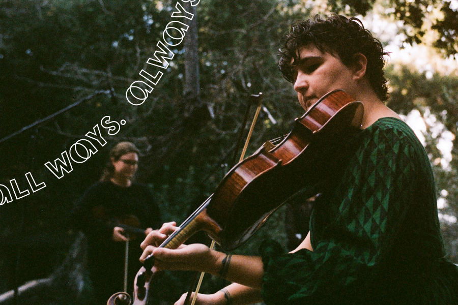 A person with medium-tone skin wearing a long forest-green garment playing the viola under trees with other musicians in the background.