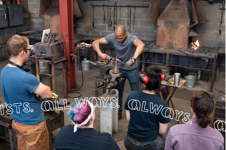 A bald Black man hammers a glowing red piece of iron around a cone in a vice as five students look on. They are standing in a blacksmithing shop filled with tools and a flaming coal forge.