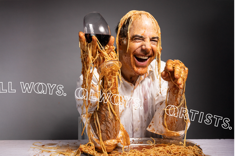 A laughing man sits at a table with a white table cloth holding a glass of red wine. He is covered in spaghetti and spaghetti sauce.