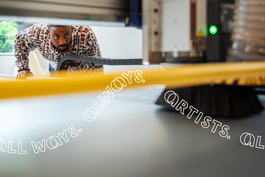 A Black man works at a large industrial machine. He is photographed through the equipment, showing his intent gaze through machinery that is out of focus in the foreground.