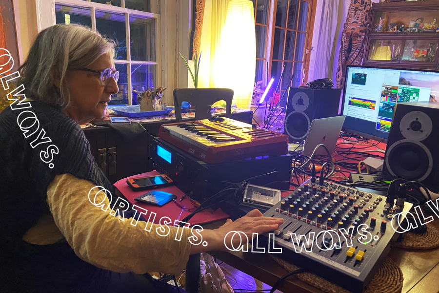 A woman with a gray bob, light complexion, and clear-colored glasses sits at a dining room table covered in sound equipment including a small keyboard, a soundboard, two speakers, and a computer monitor.