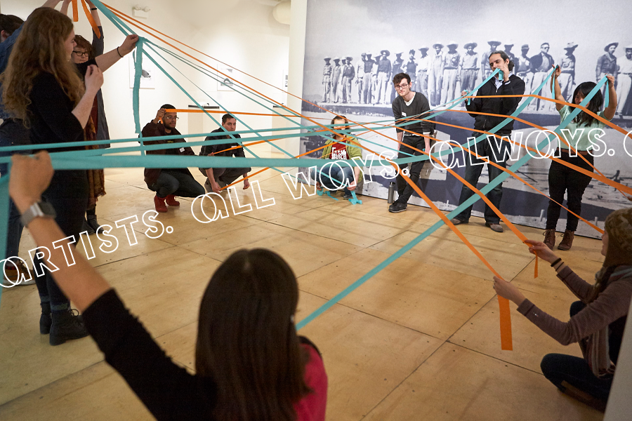 A group of people participate in an interactive activity, holding and weaving long blue-and-orange ribbons together in a web-like formation that stretches across a small gallery space. They form a circle, some standing and others crouching. Behind them is a large black-and-white mural of uniformed figures standing in a row facing us.