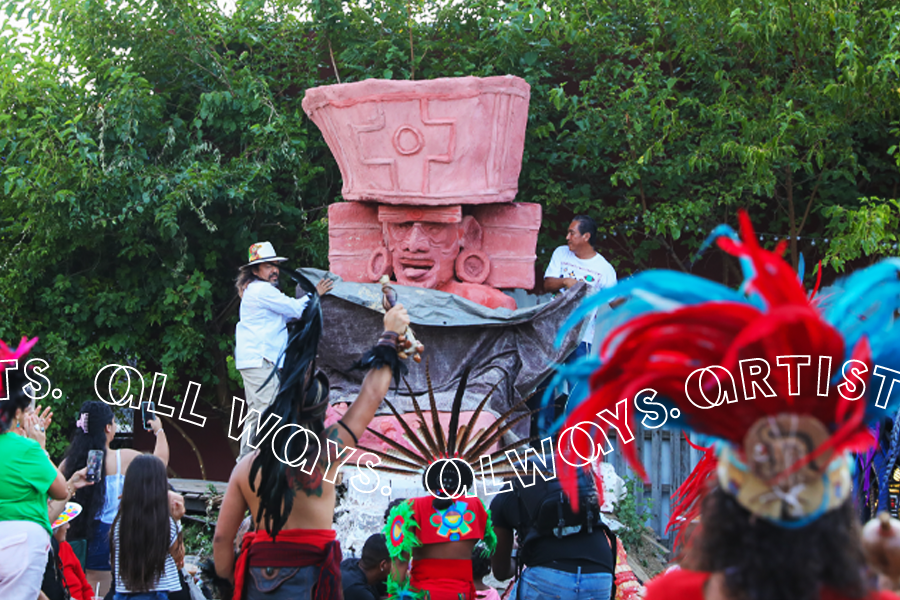 An outdoor gathering where a great terracotta colored statue of Huehueteotl, the Mesoamerican God of Fire, is being revealed from behind a gray tarp. This monumental, fifteen-foot cement figure is adorned with an enormous rhombus shaped headwear and disc-like earrings. The god opens his mouth playfully, exposing his top row of teeth and protruding tongue. The crowd surrounding the sculpture are vibrantly dressed in colorful feathers and patterns.