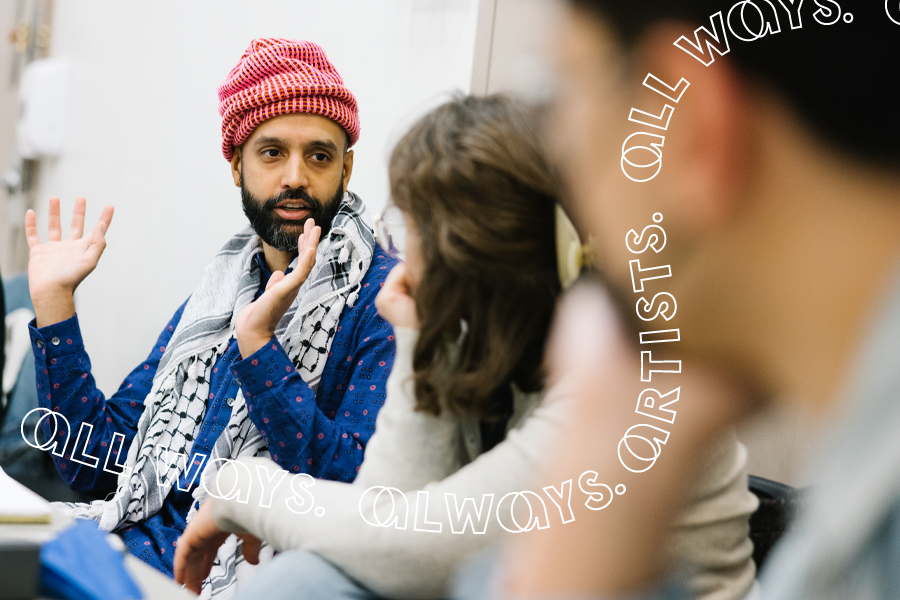 A smiling, bearded Indian man wearing a pink beanie and a blue kurta patterned with pink flowers, speaks to a rehearsal room, his hand outstretched, illustrating something. A woman sitting next to him looks at him warmly.