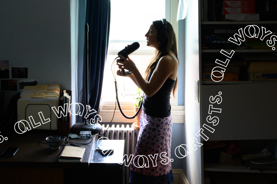 Seen in silhouette, a woman stands in a dark home office illuminated by a single window. Framed in the daylight, the woman stands in profile as she speaks into a microphone.