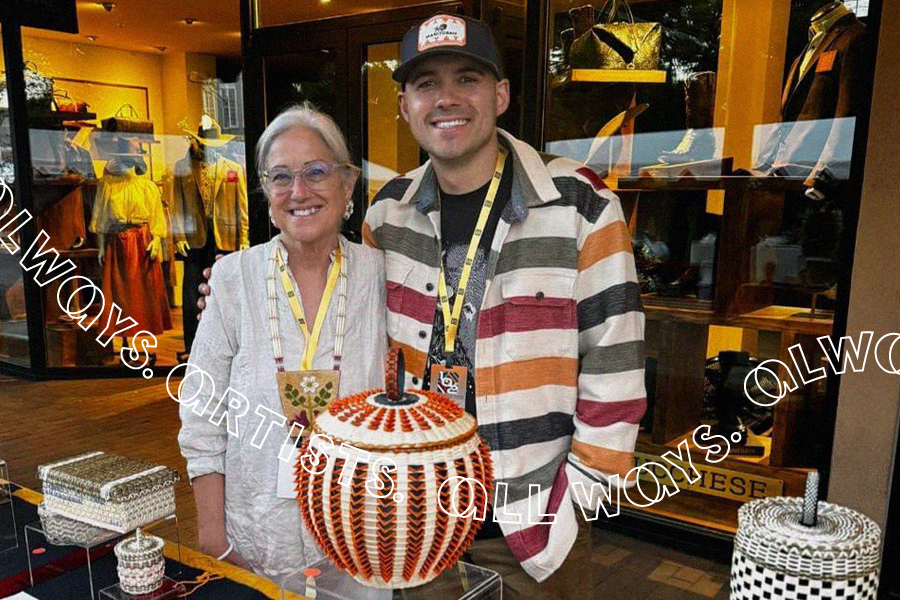 An older woman in a light-colored blouse and a younger man wearing a cap and a striped overshirt stand behind a table with baskets on it and prize ribbons below.