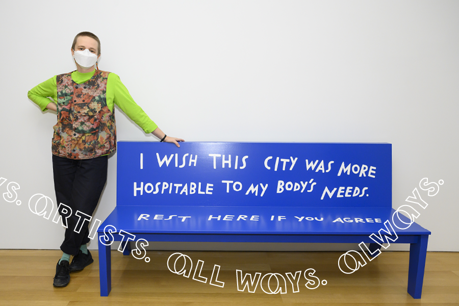 A person with a buzz cut wearing a KF94 mask, fluorescent lime shirt, and a floral fisherman-style vest poses next to a bright blue bench. Handwriting on the bench in white reads, "I wish this city was more hospitable to my body's needs. Rest here if you agree."