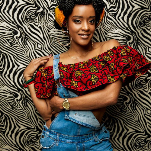 RyanNicole, an African-American woman in overalls and a patterned red top, stands smiling at the camera in front of a zebra-print background. She is posed so that her left arm wraps around her waist and her right hand rests on her shoulder.