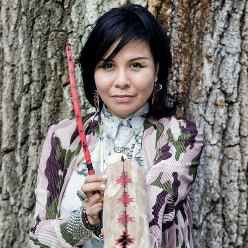 A portrait of a woman standing in front of an enormous tree holding an Apache Violin. She looks directly at the viewer, holding the instrument on her left forearm in front of her torso. She has long dark hair, wing-tipped eyeliner, and colorful patterned garments that both clash with and complement one another.