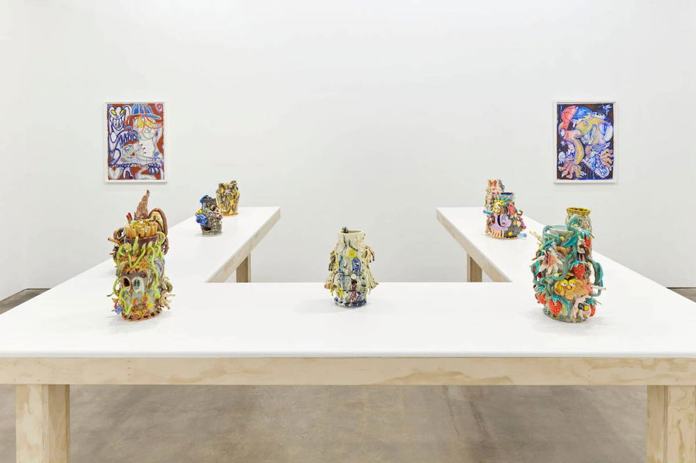 An in-gallery photograph, displaying seven sculptures atop a U-shaped table. The sculptures are vessels ranging in size and bursting with organic shapes. Many look as if they are covered in flora and fauna and others are so abstracted that the colorful shapes are unrecognizable at a distance.